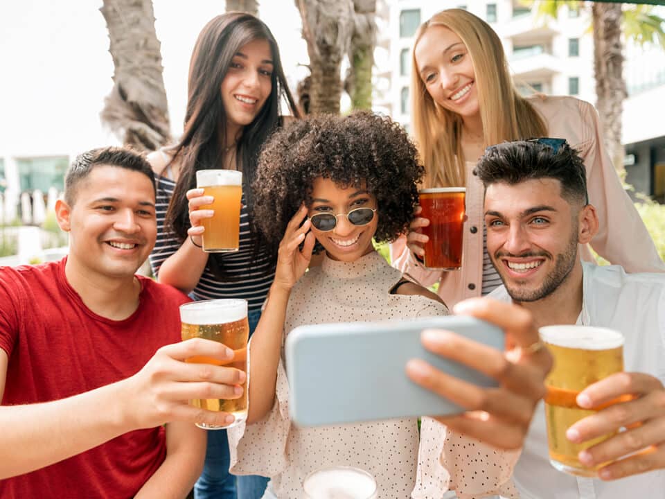 Group of friends posing for a selfie with pints of cold beer raising their glasses in a toast to the camera on the mobile phone with happy smiles